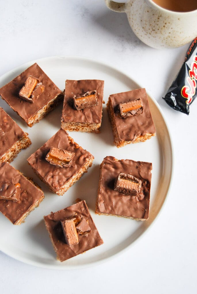 eight chocolate rice crispy squares topped with milk chocolate and topped with a Mars Bar and a large beige and white coffee mug.