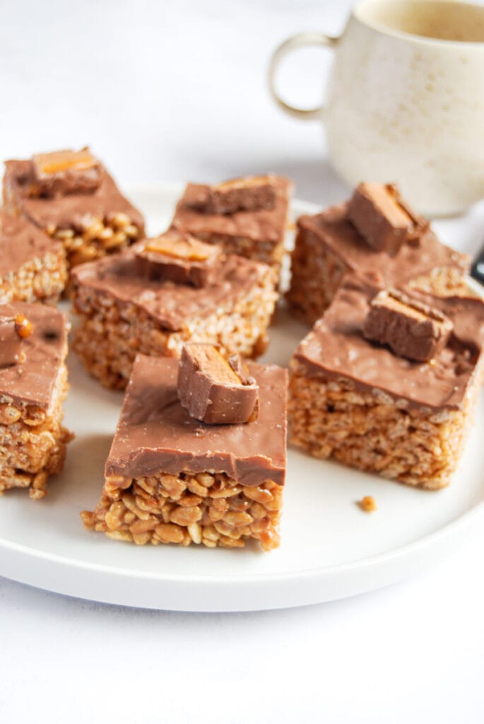 a white plate of Mars bar rice crispy squares topped with milk chocolate and a Mar Bar slice and a large beige coffee mug.