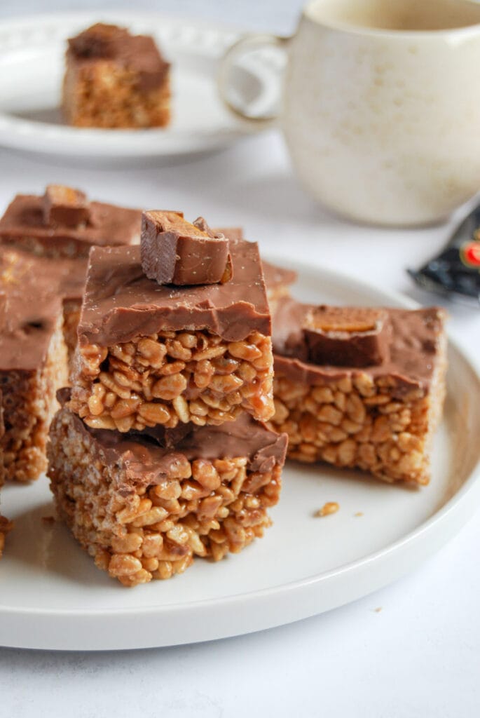 chocolate rice crispy squares topped with a Mars Bar slice on a white plate, a large coffee mug and a white plate with a chocolate crispy square.