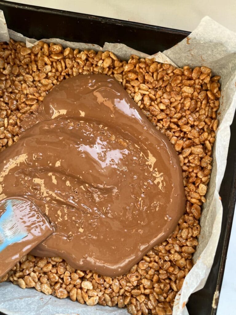 melted milk chocolate being spread over a rice crispy cake in a square baking tin.