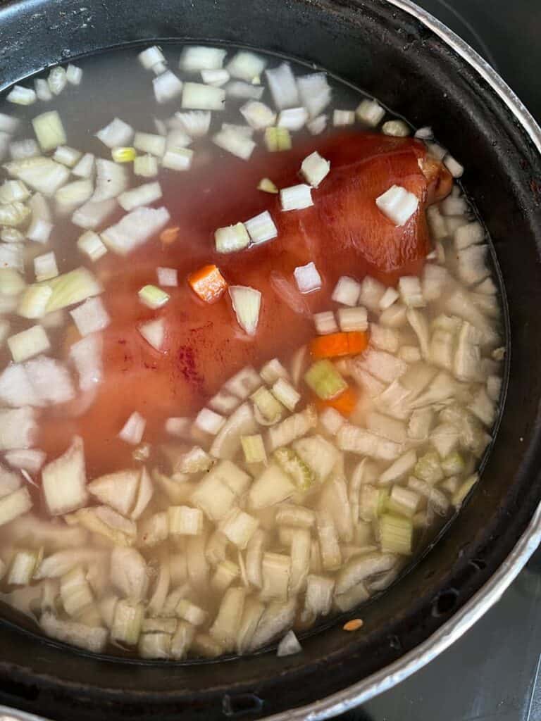 a large pan filled with water, diced onions, celery, carrots and a ham hock.