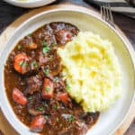 a beige bowl of beef stew in a rich Guinness gravy with carrots, parsnips and mushrooms and mashed potato with shredded cabbage. The bowl sits on a round wooden serving board. A white bowl of mash potatoes can be seen in the top left corner.