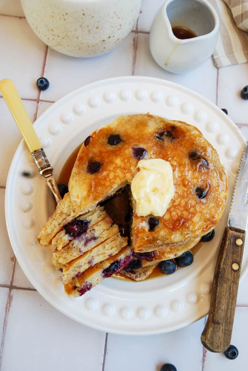 a stack of blueberry pancakes drizzled with maple syrup and topped with butter on a white plate with a fork and knife, a small white jug of maple syrup and a speckled beige and white coffee mug.