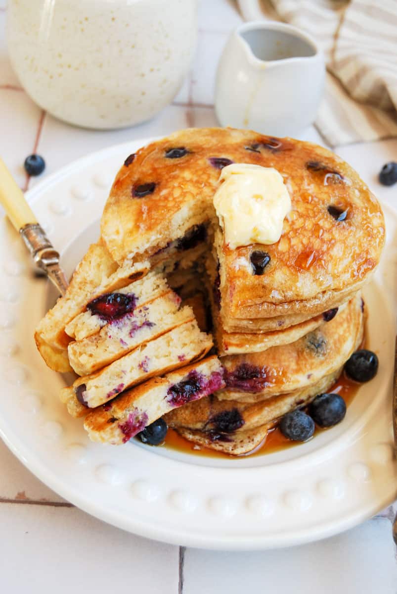 a stack of blueberry pancakes topped with butter and drizzled with maple syrup on a white plate, a small jug of maple syrup and a speckled beige and white coffee mug.