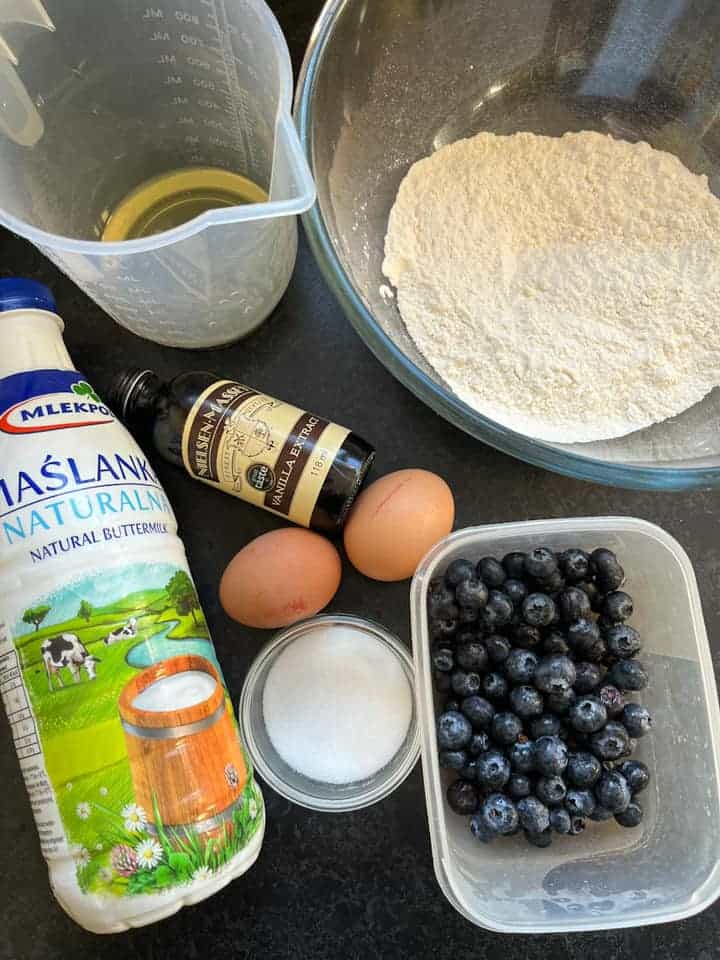 a large glass bowl of flour, a plastic measuring jug of oil, two eggs, a bottle of vanilla extract, a bottle of buttermilk, a small jar of sugar and a plastic tub of fresh blueberries.