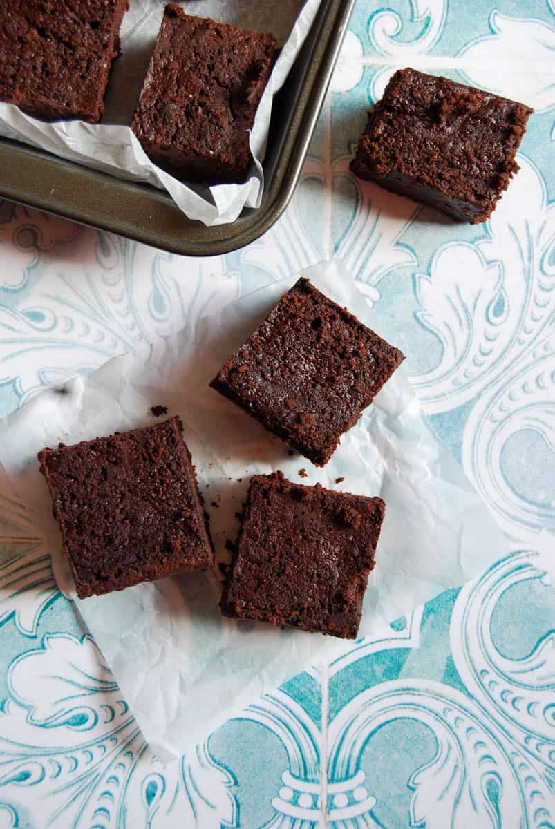 four chocolate beetroot brownies on a blue and white patterned background. A tin of brownies can be partially seen in the top left corner.