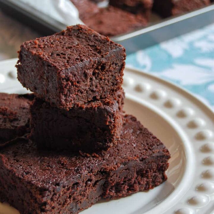 five chocolate beetroot brownies on a white plate and a tin of brownies behind the plate.