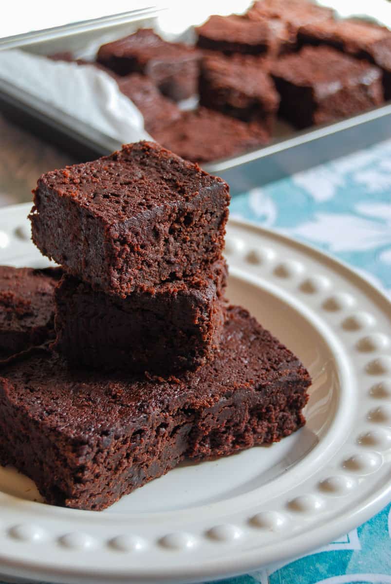 five chocolate beetroot brownies on a white plate and a tin of brownies behind the plate.