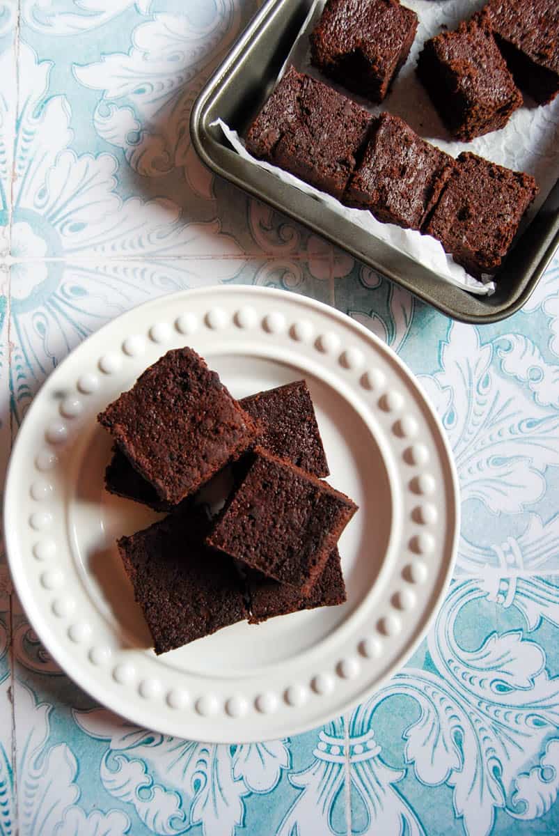 six chocolate beetroot brownies on a white textured plate and light blue and white patterned background. A tray of brownies sits above the plate.