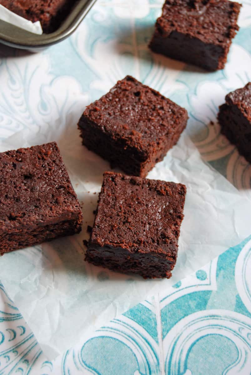 five chocolate beetroot brownies sitting on a piece of baking parchment and blue and white patterned background.