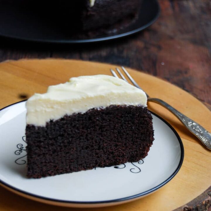A slice of chocolate Guinness cake topped with cream cheese icing on a white and black plate. The plate is sitting on a round wooden serving board with a silver fork.