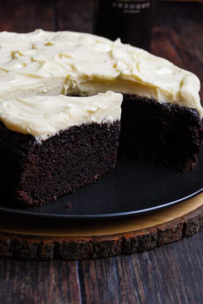 a partially sliced chocolate cake topped with cream cheese icing on a black plate. The plate is sitting on a round wooden serving board.