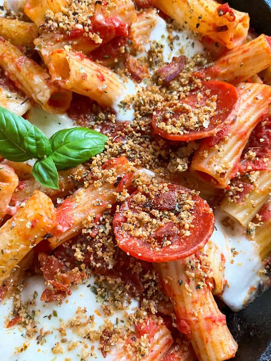 a close up picture of a pan of rigatoni pasta with tomato sauce, pepperoni slices, herb breadcrumbs and topped with a fresh basil leaf.