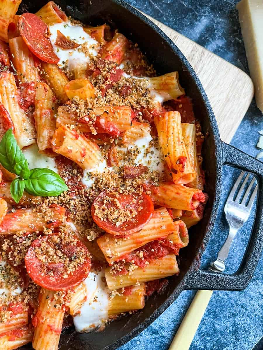 a black pan of rigatoni pasta with tomato sauce, pepperoni slices, mozzarella cheese topped with herby breadcrumbs and a fresh basil leaf.