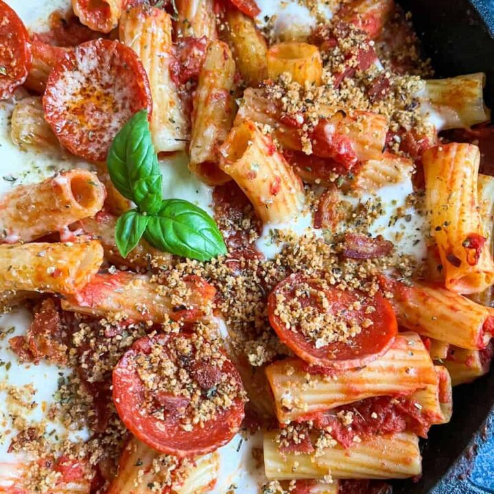 a black pan of rigatoni pasta with pepperoni slices, mozzarella cheese and topped with herb breadcrumbs and a basil leaf.