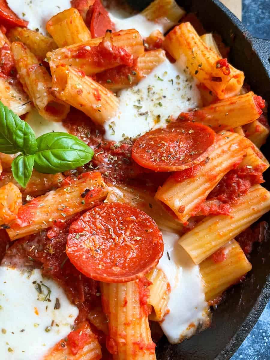 A close up image of a pan of pasta in tomato sauce with sliced pepperoni, mozarella cheese and a fresh basil leaf. 