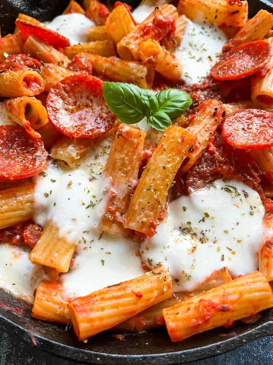 a close up photo of a pan of rigatoni pasta in tomato sauce, mozzarella cheese, pepperoni slices topped with dried oregano and a fresh basil leaf.