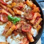 a black saucepan of rigatoni pasta in tomato sauce with pepperoni slices, mozzarella cheese sprinkled with crispy herb breadcrumbs and a fresh basil leaf.