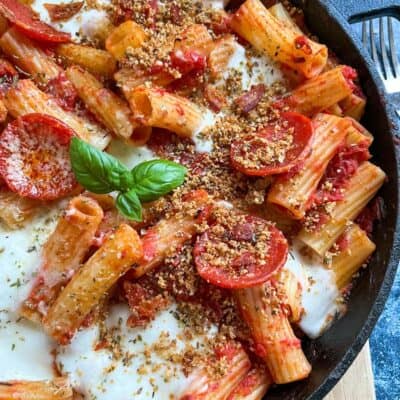 a black saucepan of rigatoni pasta in tomato sauce with pepperoni slices, mozzarella cheese sprinkled with crispy herb breadcrumbs and a fresh basil leaf.