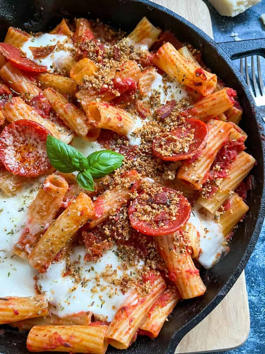 a black saucepan of rigatoni pasta in tomato sauce with pepperoni slices, mozzarella cheese sprinkled with crispy herb breadcrumbs and a fresh basil leaf.