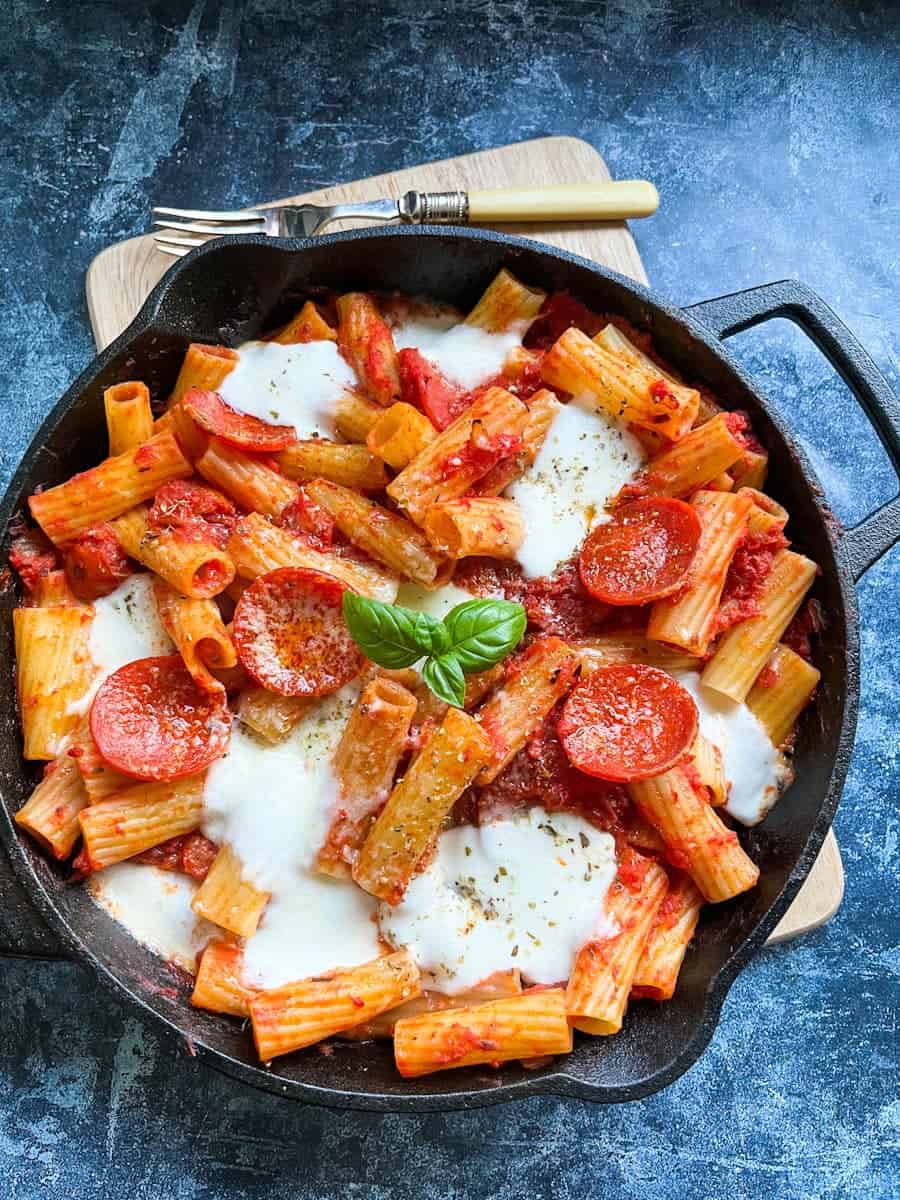 a black skillet pan of rigatoni pasta in tomato sauce topped with pepperoni slices, mozzarella cheese and a fresh basil leaf.