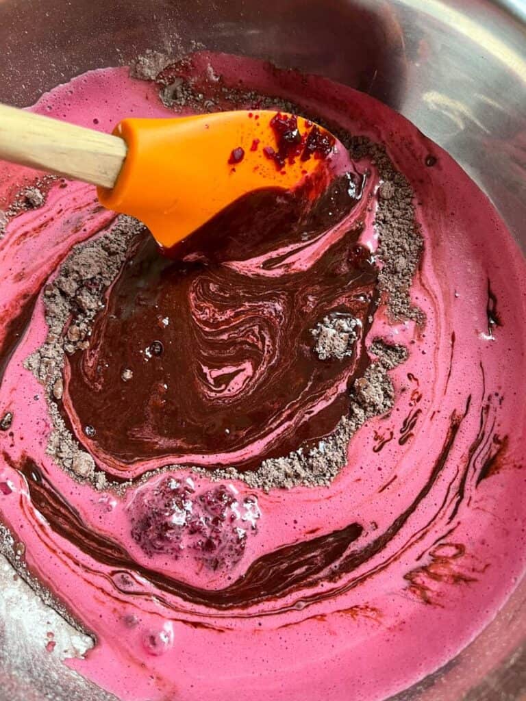 pureed beetroot, eggs and sugar being stirred in a silver bowl of cocoa powder and plain flour with an orange wooden handled spatula.