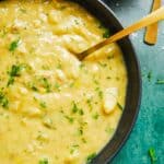 A black shallow bowl of leek and potato soup with bacon and scattered with chopped parsley and chives with a gold spoon.