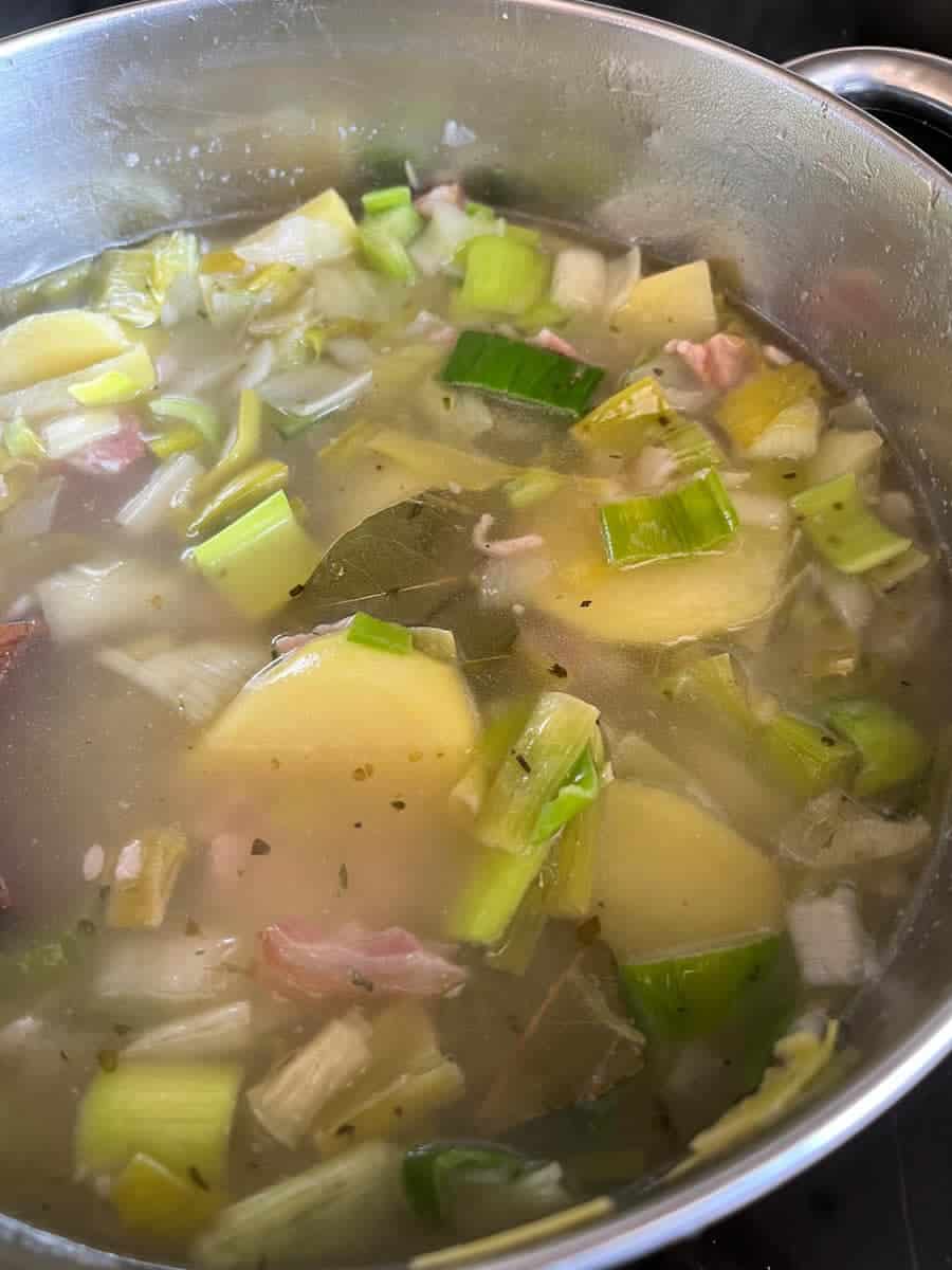 a silver saucepan of water with diced potatoes, sliced leeks, bacon, herbs and a bay leaf .