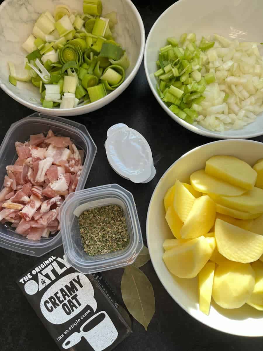 white bowls of sliced potatoes, onion, celery, leeks, a small plastic dish of bacon, a pot of dried herbs, two bay leaves, a chicken stock pot and a cardboard container of oat cream.