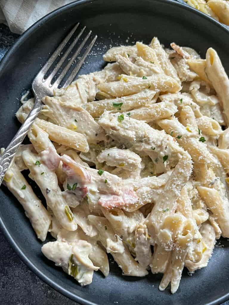 a black bowl of chicken pasta with bacon and leeks in a creamy sauce with a silver fork.