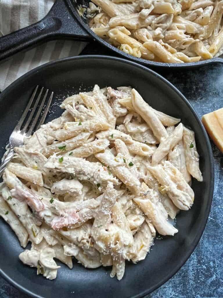 a black stoneware dish with penne pasta, chicken and bacon in a cream sauce sprinkled with parmesan cheese.