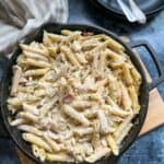 a pan of creamy chicken pasta with leeks and bacon sprinkled with parmesan cheese on a wooden serving board.