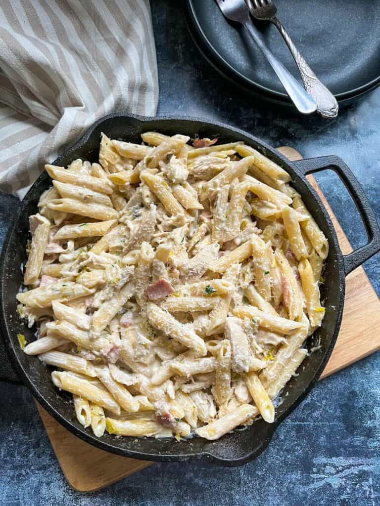 a pan of creamy chicken pasta with leeks and bacon sprinkled with parmesan cheese on a wooden serving board.