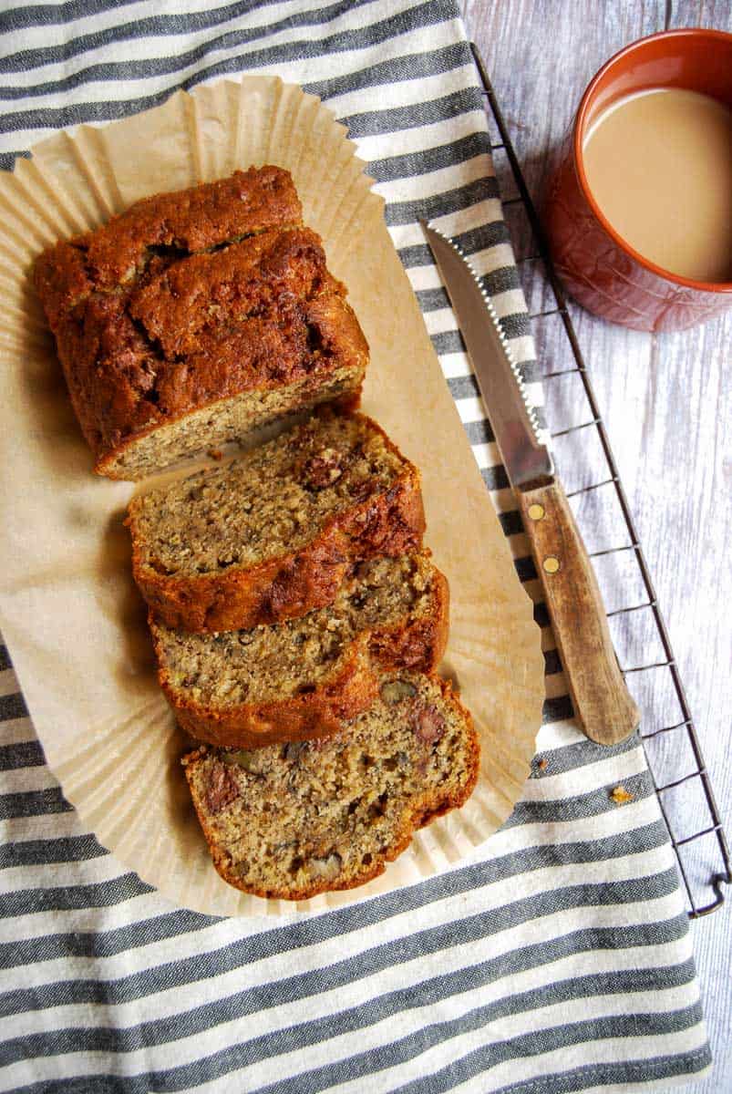 A sliced banana bread on a piece of baking paper sitting on a cooling rack covered with a striped tea towel, a wooden handled knife and a cup of coffee.