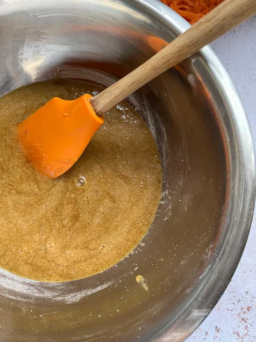 a silver bowl of beaten eggs, brown sugar and oil and a wooden handled orange spatula.