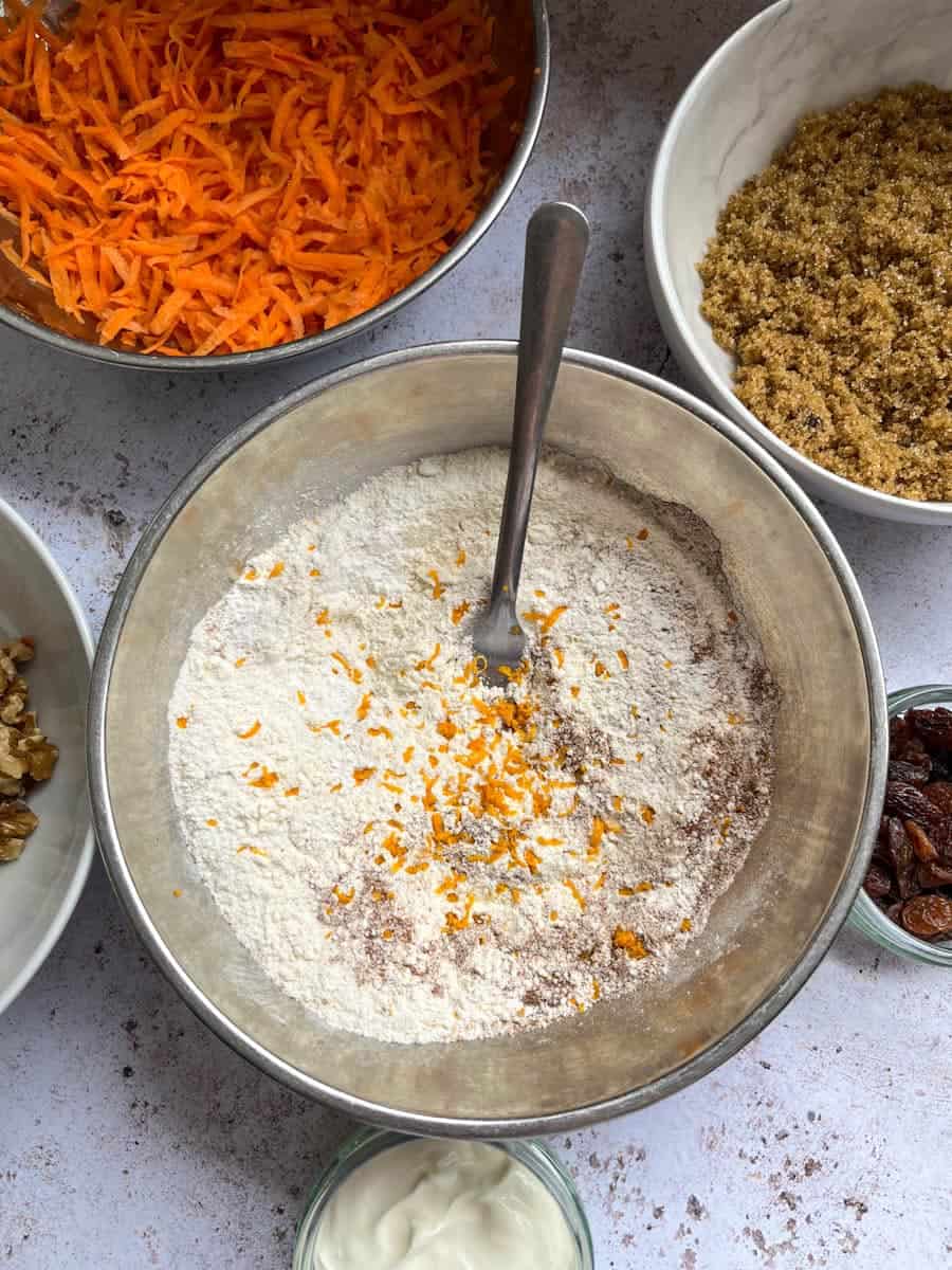 a silver bowl of plain flour and grated orange zest with a silver fork, a silver bowl of grated carrots, a white bowl of brown sugar, a small dish of sultanas and a dish of sour cream.