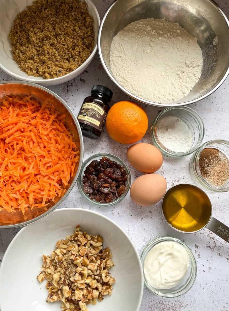 Bowls of flour, brown sugar, grated carrot, walnuts and small dishes of spices, baking powder, sour cream, sultanas, oil, two eggs, a bottle of vanilla extract and an orange.