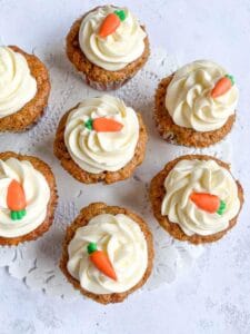 seven carrot cupcakes topped with a cream cheese icing swirl and a miniature fondant carrot decoration on a white lace paper doily.