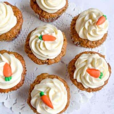 seven carrot cupcakes topped with a cream cheese icing swirl and a miniature fondant carrot decoration on a white lace paper doily.