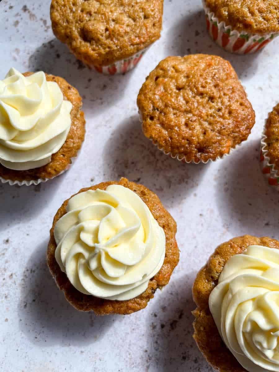 six carrot cake cupcakes on a white and grey background. Three cakes have been decorated with a cream cheese icing swirl.