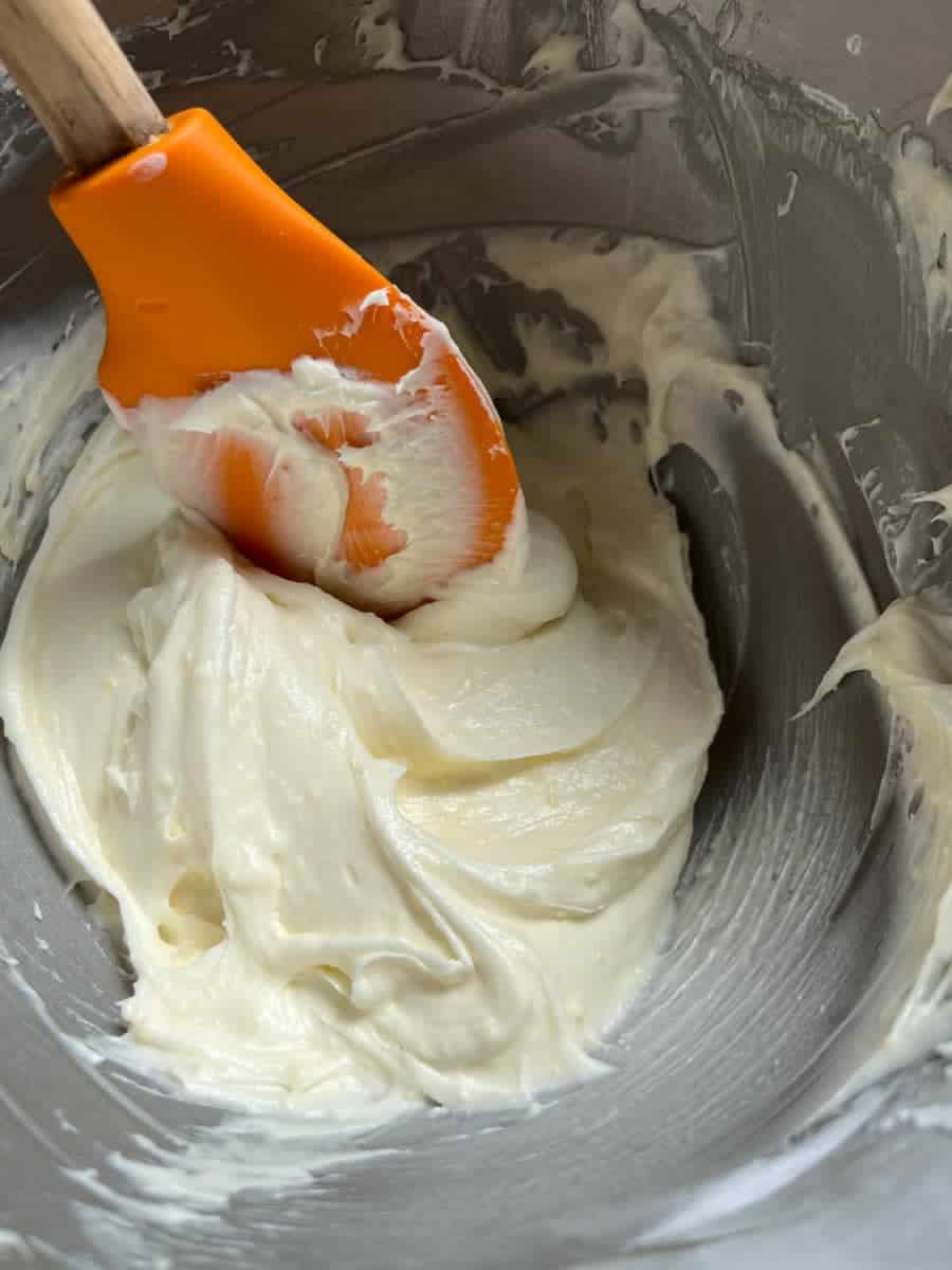 a silver bowl of cream cheese icing with an wooden handled orange spatula.