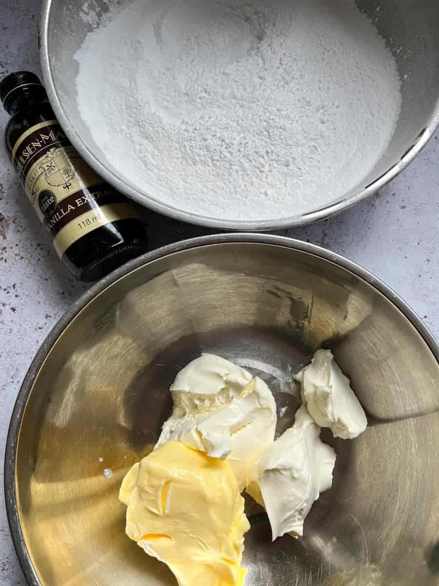 a silver bowl of icing sugar, a silver bowl of soft butter and cream cheese and a bottle of vanilla extract.