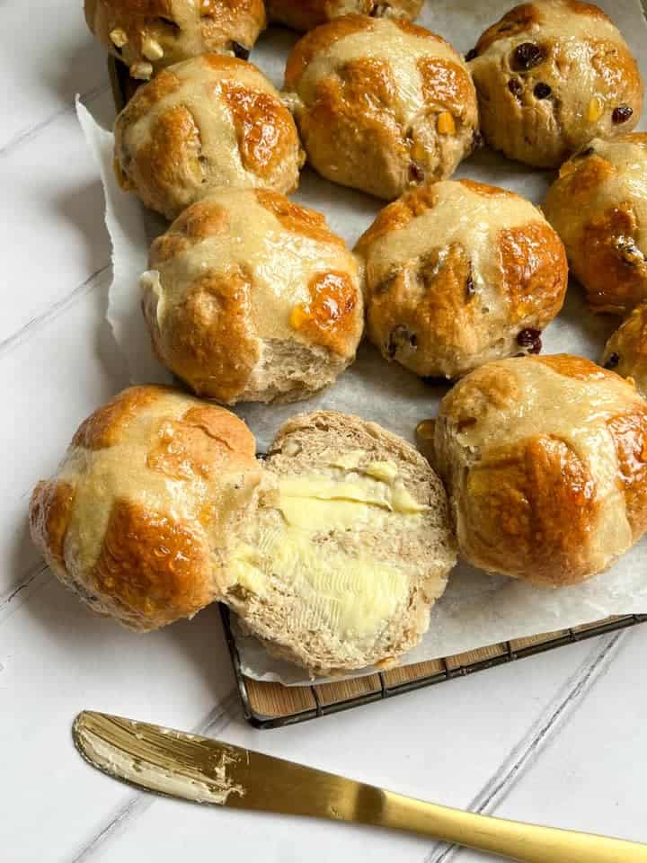 a batch of hot cross buns on a wire cooling rack lined with baking parchment. One of the buns has been split open and buttered.