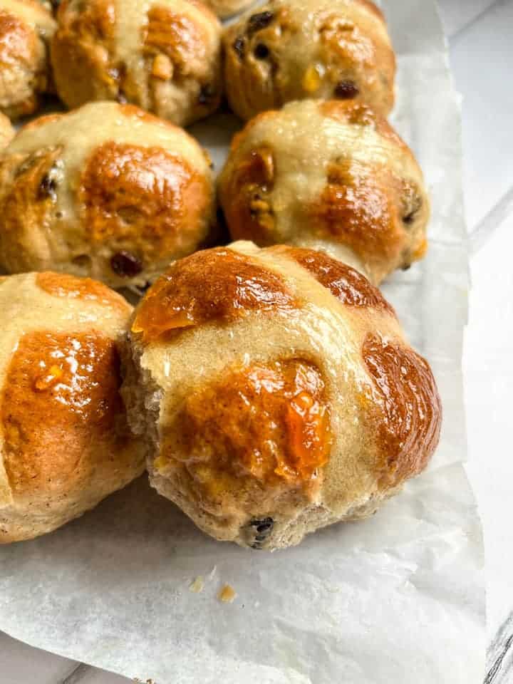 A batch of hot cross buns brushed with orange marmalade on a wire rack lined with baking parchment.