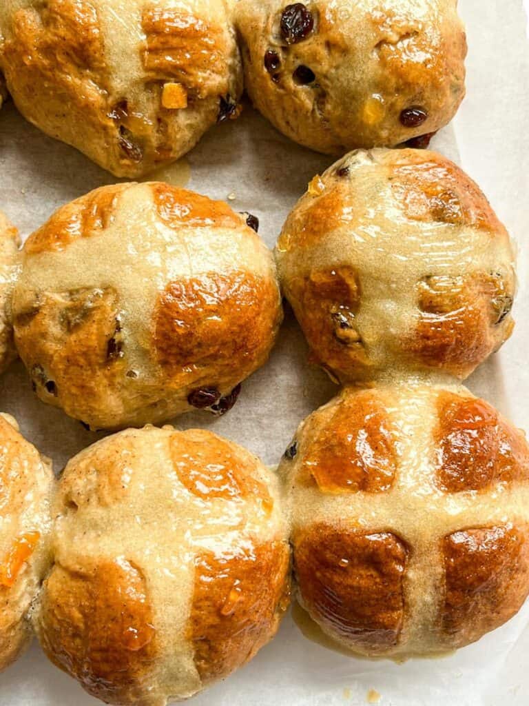 a batch of hot cross buns brushed with orange marmalade on a piece of baking parchment.