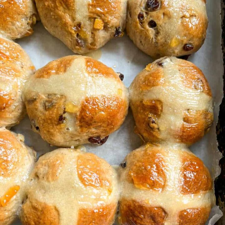 a batch of hot cross buns glazed with orange marmalade on a baking tray lined with parchment.