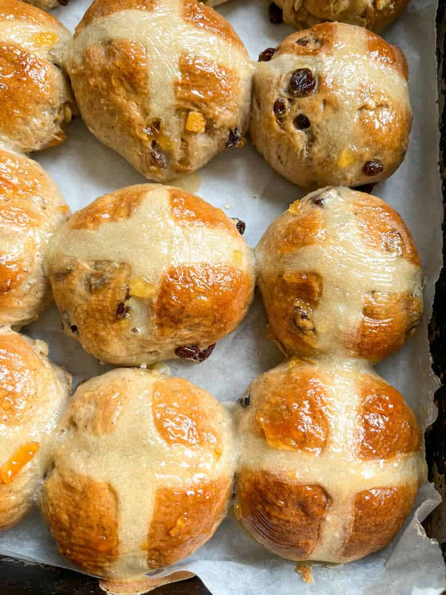 a batch of hot cross buns glazed with orange marmalade on a baking tray lined with parchment.