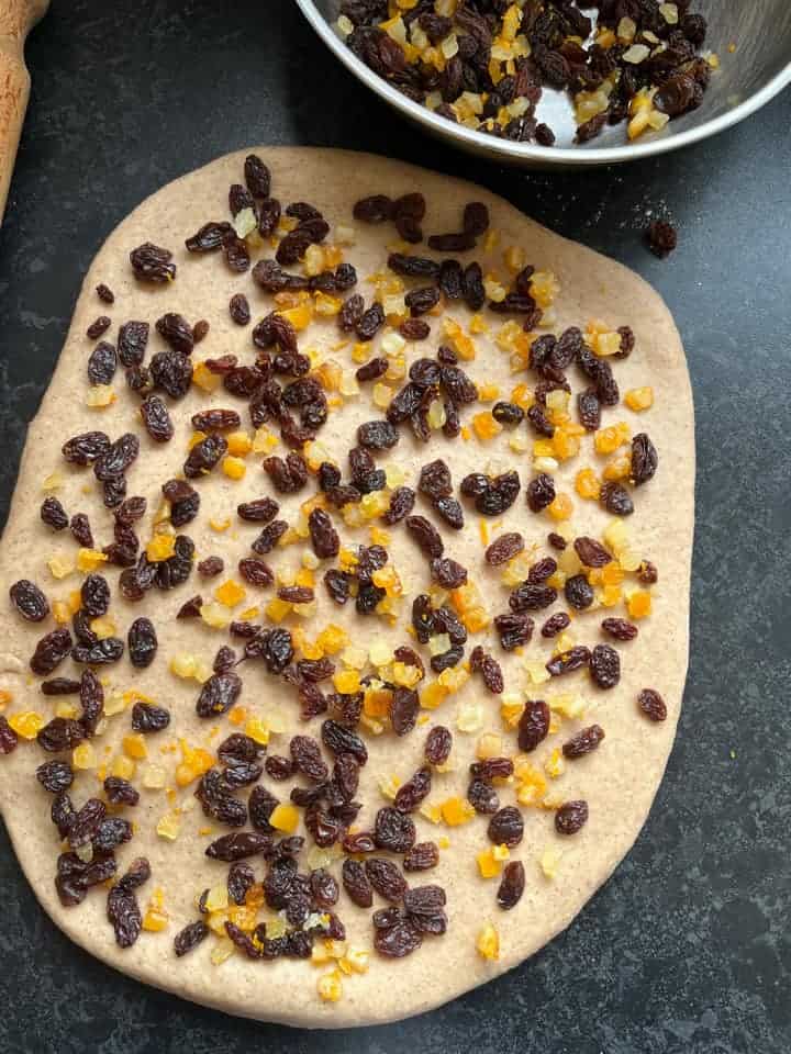 a large piece of bread dough scattered with raisins and candied mixed peel and a silver bowl of dried fruit on a dark grey work surface.