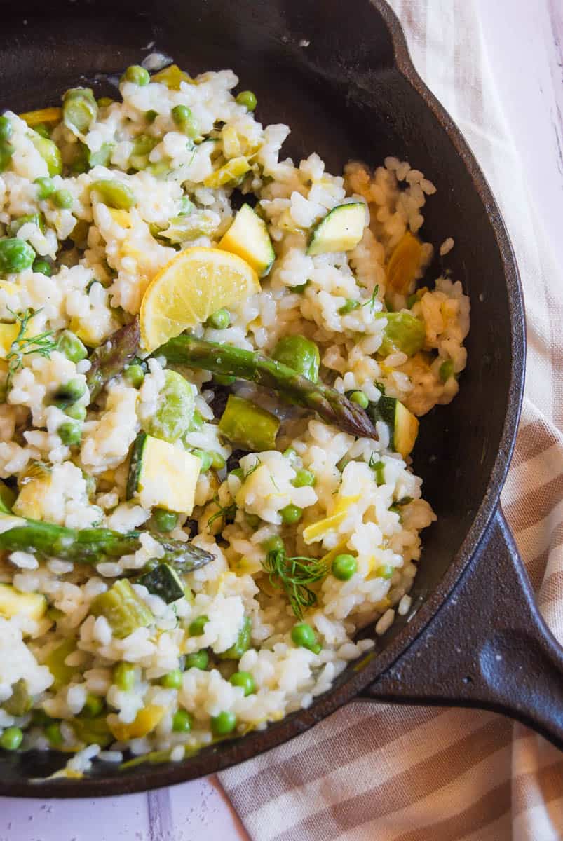 a black cast iron dish of risotto with asparagus, sliced leeks, peas, broad beans and a slice of lemon resting on a white and beige striped tea towel.