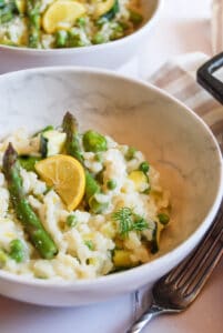 two bowls of risotto with asparagus, broad beans, peas, fresh dill and a slice of lemon. A silver fork sits beside the bowl at the front.